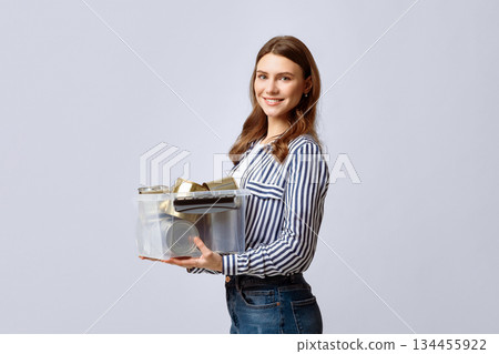 A young woman stands indoors, smiling broadly while holding a transparent container filled with recyclable materials. She showcases a commitment to eco-friendly practices and sustainability. A young woman stands indoors, smiling broadly while holding a transparent container filled with recyclable materials. She showcases a commitment to eco-friendly practices and sustainability. 134455922
