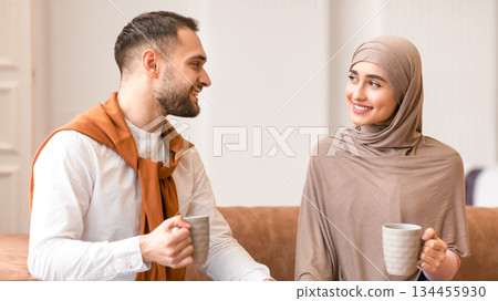Young Muslim Couple Talking And Flirting Drinking Coffee Enjoying Conversation Sitting On Couch Smiling Each Other At Home. Beautiful Arab Woman Touching Husband's Hand. Love, Romantic Relationship 134455930
