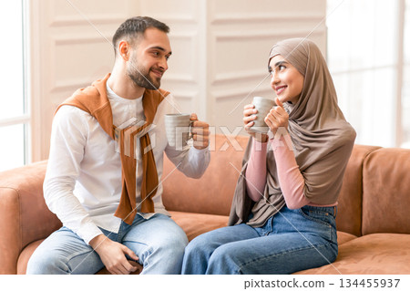 Romantic Relationship. Cheerful Muslim Spouses Drinking Coffee Sitting On Couch Smiling Each Other Posing At Home. Arabic Young Couple Flirting And Bonding Spending Time Together On Weekend Indoors. 134455937