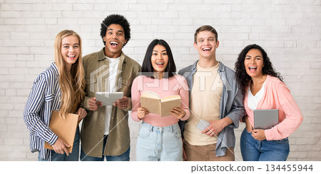 Facial expressions and emotions of student team. Excited laughing young multiracial people with open mouths hold books and gadgets and look at camera on white wall background, studio shot, empty space Facial expressions and emotions of student team. Excited laughing young multiracial people with open mouths hold books and gadgets and look at camera on white wall background, studio shot, empty space 134455944
