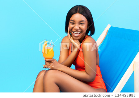 Pretty black woman drinking tropical cocktail in lounge chair, laughing, wearing stylish bikini over blue background. Young African American lady chilling on beach during summer holidays Pretty black woman drinking tropical cocktail in lounge chair, laughing, wearing stylish bikini over blue background. Young African American lady chilling on beach during summer holidays 134455995