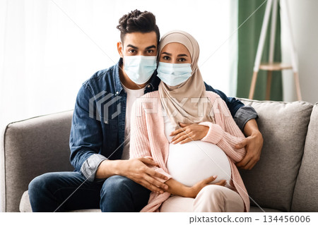 Young pregnant muslim couple wearing protective medical masks sitting on couch and looking at camera. Islamic couple awaiting baby during coronavirus pandemic, sitting on self-isolation at home 134456006