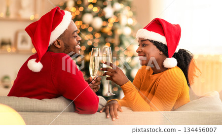 Romantic celebration of Christmas for two during coronavirus. Millennial happy african american man and lady in Santa hats clink with glasses of champagne, sit on sofa in home with tree and garlands Romantic celebration of Christmas for two during coronavirus. Millennial happy african american man and lady in Santa hats clink with glasses of champagne, sit on sofa in home with tree and garlands 134456007