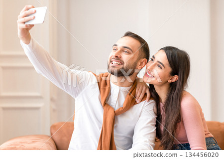 Cheerful Arab Couple Making Selfie On Mobile Phone Sitting On Sofa At Home. Modern Middle Eastern Millennials Having Fun Posing Together And Taking Photos Via Cellphone Indoor. People And Gadgets Cheerful Arab Couple Making Selfie On Mobile Phone Sitting On Sofa At Home. Modern Middle Eastern Millennials Having Fun Posing Together And Taking Photos Via Cellphone Indoor. People And Gadgets 134456020