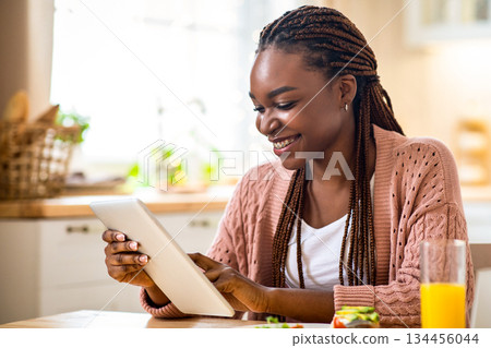 Young Smiling Black Woman Using Digital Tablet During Breakfast In Kitchen. Happy African American Lady Eating Tasty Food And Browsing Internet, Shopping Online Or Checking Social Networks, Free Space 134456044