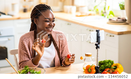 Online Nutritionist. Cheerful Young Black Female Recording Food Review In Kitchen, Demonstrating Fresh Ripe Cherry Tomatoes At Camera, Using Smartphone On Tripod For Video Content Creation, Closeup Online Nutritionist. Cheerful Young Black Female Recording Food Review In Kitchen, Demonstrating Fresh Ripe Cherry Tomatoes At Camera, Using Smartphone On Tripod For Video Content Creation, Closeup 134456052