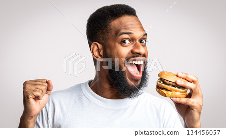 Joyful Black Guy Biting Burger Enjoying Cheat Meal Eating Unhealthy Cheeseburger Standing Posing Over White Studio Background. Junk Food Overeating, Obesity Problem And Unhealthy Nutrition Habit 134456057