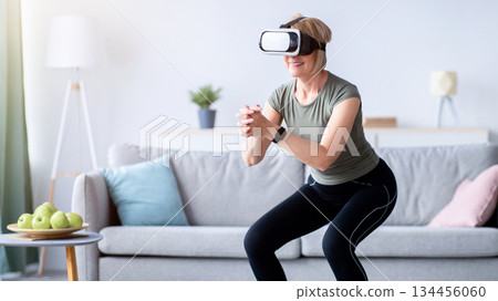 Exercising with virtual reality concept. Athletic mature woman in VR headset doing squats on her home workout. Positive senior lady using modern technologies for domestic training 134456060