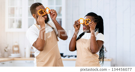 Cooking Fun. Cheerful African American Spouses In Aprons Fooling In Kitchen, Funny Black Couple Holding Bell Pepper Slices Near Eyes And Laughing, Enjoying Preparing Food Together At Home, Free Space 134456077