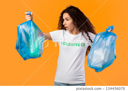 Shocked Female Volunteer Holding Unsorted Plastic Garbage Bags With Waste And Litter Standing Over Yellow Studio Background. Junk Sorting And Pollution Problem Concept 134456078