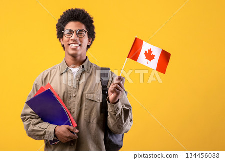 Education, school, college and university. Happy young african american guy in glasses holding notepads and small Canada flag and ready to study, isolated on orange background, free space, studio shot 134456088