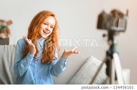 Blogging Concept. Smiling red-haired teen girl recording lifestyle blog talking to camera on tripod, sitting on the couch at home. Happy young influencer filming review for her internet channel Blogging Concept. Smiling red-haired teen girl recording lifestyle blog talking to camera on tripod, sitting on the couch at home. Happy young influencer filming review for her internet channel 134456101