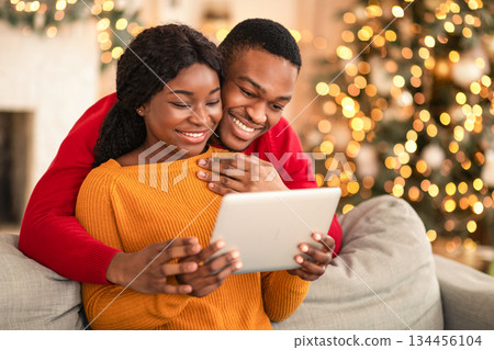 Video call on eve of Christmas. Family happy millennial african american husband and wife look at tablet and congratulate friends in cozy living room interior with Xmas tree and glowing garlands Video call on eve of Christmas. Family happy millennial african american husband and wife look at tablet and congratulate friends in cozy living room interior with Xmas tree and glowing garlands 134456104