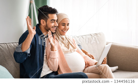 Online Communication. Cheerful Pregnant Middle Eastern Spouses Making Video Call With Digital Tablet While Relaxing Together On Couch In Living Room, Happy Couple Waving Hands At Camera And Smiling Online Communication. Cheerful Pregnant Middle Eastern Spouses Making Video Call With Digital Tablet While Relaxing Together On Couch In Living Room, Happy Couple Waving Hands At Camera And Smiling 134456116