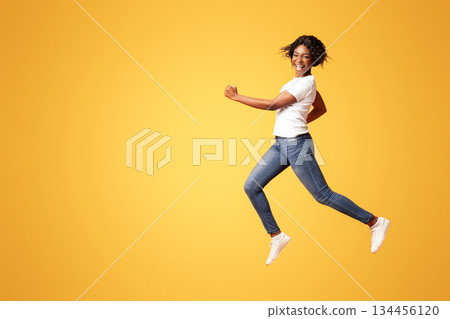 Active black lady in casual outfit running over orange studio background, full length shot. Cheerful african american young woman in jeans and white t-shirt having fun on her own 134456120