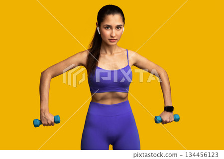 Sport And Fitness Concept. Portrait Of Confident Young Strong Woman In Wireless Headphones Exercising With Two Blue Dumbbells, Listening To Music, Posing Isolated On Orange Studio Background Sport And Fitness Concept. Portrait Of Confident Young Strong Woman In Wireless Headphones Exercising With Two Blue Dumbbells, Listening To Music, Posing Isolated On Orange Studio Background 134456123