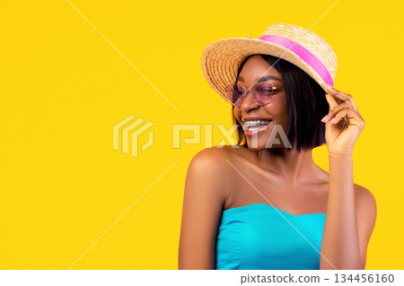 Portrait of cheerful black lady with dental braces wearing summer dress, sunglasses and straw hat on orange studio background. Happy African American posing in summertime outfit 134456160