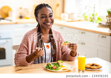 Cheerful Black Lady Eating Tasty Breakfast Or Lunch In Kitchen At Home. Joyful Young African American Female Sitting At Table Enjoying Her Delicious Meal And Drinking Orange Juice, Copy Space 134456191