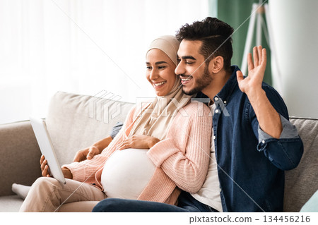 Joyful Expectant Muslim Spouses Making Video Call With Digital Tablet At Home, Happy Pregnant Islamic Couple Talking With Friends Or Family Via Web Conference, Waving Hand At Camera And Smiling 134456216