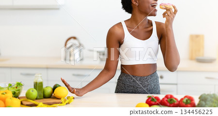 Black Fitness Female Enjoying Cheat Meal Eating Donut Posing In Kitchen At Home. Sugary Food And Desserts, Bad Nutrition Habit, Junk Food Craving And Overeating Concept 134456225