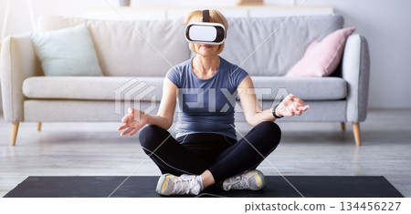 Full length of senior lady in VR glasses practicing yoga or meditating in lotus pose at living room. Mature woman exploring virtual reality sports, exercising her mindfulness at home Full length of senior lady in VR glasses practicing yoga or meditating in lotus pose at living room. Mature woman exploring virtual reality sports, exercising her mindfulness at home 134456227