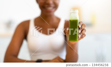 Slim African Female Showing Green Smoothie Bottle Smiling To Camera Posing Sitting In Modern Kitchen Indoors. Healthy Detox Diet For Weight Loss. Vegetable Juice And Drinks Recipes For Slimming 134456238