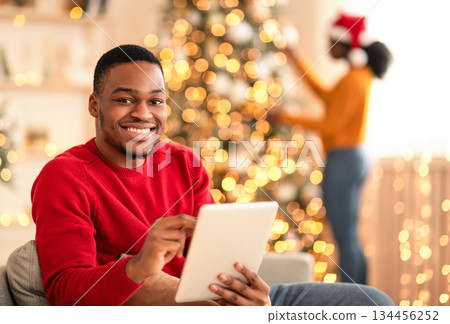 Christmas eve shopping online during sale and discount, and video call to family. Smiling millennial african american man typing on tablet, woman decorating tree in living room interior to garlands Christmas eve shopping online during sale and discount, and video call to family. Smiling millennial african american man typing on tablet, woman decorating tree in living room interior to garlands 134456252