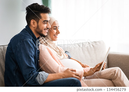 Pregnant Muslim Woman And Her Husband Relaxing With Digital Tablet At Home, Happy Islamic Spouses Shopping Online In Internet Or Browsing Social Networks, Enjoying Spending Time Together, Free Space Pregnant Muslim Woman And Her Husband Relaxing With Digital Tablet At Home, Happy Islamic Spouses Shopping Online In Internet Or Browsing Social Networks, Enjoying Spending Time Together, Free Space 134456279
