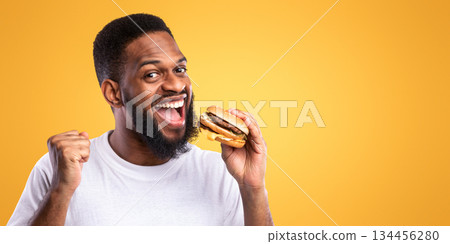 Excited African Man Eating Burger Gesturing Yes Enjoying Unhealthy Fast Food Standing Over Yellow Studio Background. Finally, Cheat Meal. Tasty Junk Food Concept Excited African Man Eating Burger Gesturing Yes Enjoying Unhealthy Fast Food Standing Over Yellow Studio Background. Finally, Cheat Meal. Tasty Junk Food Concept 134456280