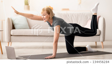 Home sports. Fit senior woman working out in front of laptop, lifting her leg and arm in living room. Athletic mature lady exercising on yoga mat, doing domestic fitness to online training program 134456284