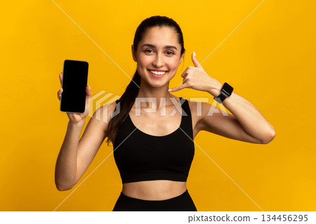 Portrait of smiling fit young lady holding smartphone with black empty screen for mockup template, showing call me back gesture, wearing fitness tracker, orange studio background 134456295
