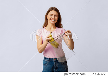 Smiling Young Lady Using Reusable String Mesh Bag For Grocery Shopping, Holding Fresh Organic Bananas And Smiling At Camera, Eco-Friendly Woman Posing Over Light Studio Background, Copy Space 134456327