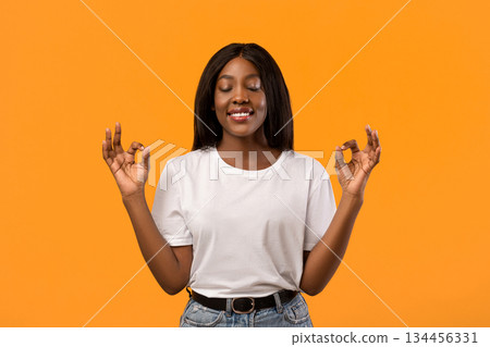 Staying calm and relaxed in urban atmosphere. Smiling african american woman in casual outfit, closing eyes and raising hands in zen gesture, meditating or practicing yoga over orange background 134456331
