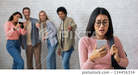 Humiliation, education and bullying. Sad asian young female in glasses looks at phone, zoomers multi ethnic students make video and laugh at lady, on white brick wall, copy space, studio shot Humiliation, education and bullying. Sad asian young female in glasses looks at phone, zoomers multi ethnic students make video and laugh at lady, on white brick wall, copy space, studio shot 134456334