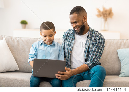 Black Father And Son Using Laptop Computer Watching Cartoons Online Sitting On Sofa Indoors On Weekend. Young Dad And His Kid Browsing Internet Together. Modern Gadgets Concept Black Father And Son Using Laptop Computer Watching Cartoons Online Sitting On Sofa Indoors On Weekend. Young Dad And His Kid Browsing Internet Together. Modern Gadgets Concept 134456344