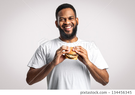 Junk Food. Happy Black Man Holding Burger Standing On White Background In Studio, Smiling Looking At Camera. Nutrition, Overeating Habit And Unhealthy Eating Concept Junk Food. Happy Black Man Holding Burger Standing On White Background In Studio, Smiling Looking At Camera. Nutrition, Overeating Habit And Unhealthy Eating Concept 134456345