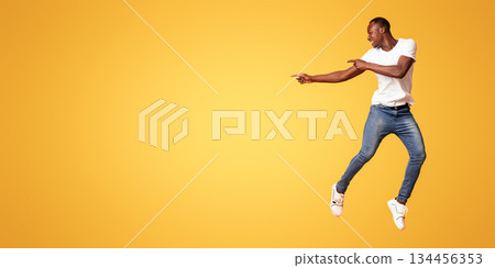 Stylish millennial african american man pointing at copy space while jumping up over orange studio background. Happy black guy pointing at empty space for text or advertisement, full length shot 134456353