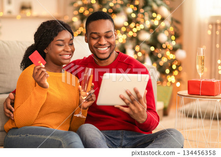 Buying gifts and online shopping before holidays. Cheerful millennial husband and wife look at tablet and credit card in cozy living room interior with Christmas tree and glowing garlands, copy space 134456358