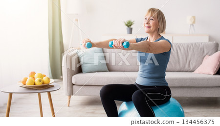 Strong mature woman working out with dumbbells on fitness ball at home, blank space. Fit senior lady taking care of her health, having domestic training, leading active lifesyle, indoors 134456375