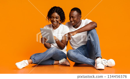 Smiling african american lovers sitting together on floor and using digital tablet over orange studio background, copy space. Young black woman and man using new application on modern digital pad 134456376