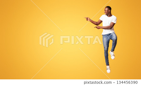 Excited black guy jumping up and showing copy space over orange studio background. Emotional african american millennial handsome man smiling and pointing at advertising or announcement 134456390