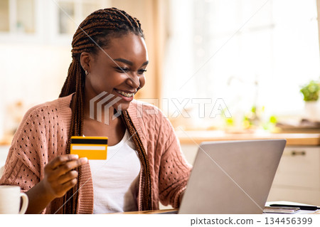 Online Shopping. Positive Black Lady Using Credit Card And Laptop Computer In Kitchen, Smiling African American Woman Purchasing Goods In Internet Or Making Easy Utility Payments, Free Space 134456399
