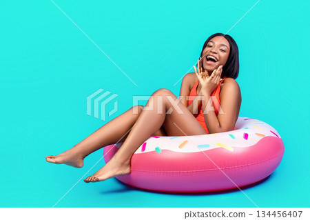 Portrait of excited black lady in bikini touching her face, sitting in donut shaped inflatable ring on blue studio background. Full length of emotional young woman enjoying beach party 134456407