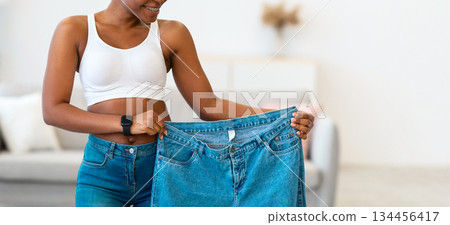 Joy Of Weight Loss. Black Lady Holding Oversized Jeans Too Big After Great Weight-Loss And Successful Slimming Standing Posing At Home. Dieting Motivation Concept 134456417