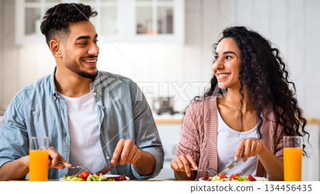 Romantic middle-eastern couple having healthy breakfast together in kitchen at home, happy arab man and woman eating delecious meal, drinking orange juice and looking at each other, closeup 134456435