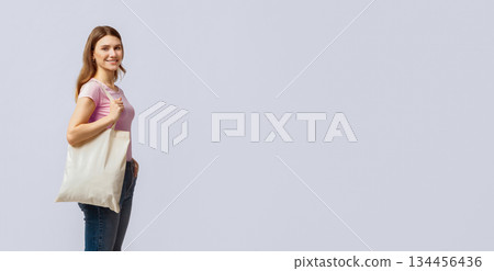 A young woman holds a reusable tote bag as she promotes recycling practices. She stands confidently against a neutral background, encouraging others to adopt eco-friendly habits, copy space 134456436