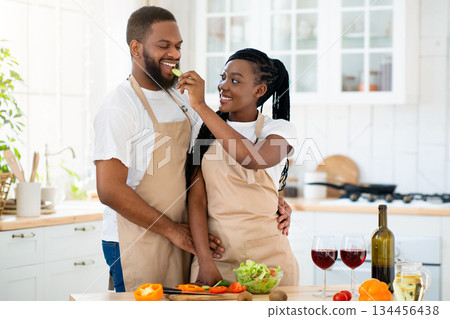 Cheerful black couple cooking and tasting healthy food in kitchen at home. Loving woman feeding her man with cucumber while they preparing tasty meal and drinking wine together, free space 134456438