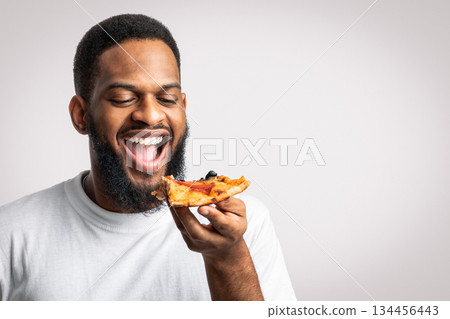 Joyful African Millennial Guy Eating Pizza Slice Posing With Opened Mouth Standing Over White Background In Studio. Junk Food Eater Enjoying Pizza. Pizzeria Delivery Concept Joyful African Millennial Guy Eating Pizza Slice Posing With Opened Mouth Standing Over White Background In Studio. Junk Food Eater Enjoying Pizza. Pizzeria Delivery Concept 134456443