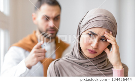 Aggressive Muslim Husband Shouting At Depressed Wife Tired Of Domestic Violence And Victimization Standing At Home. Selective Focus. Relationship With Abuser And Family Conflicts 134456444