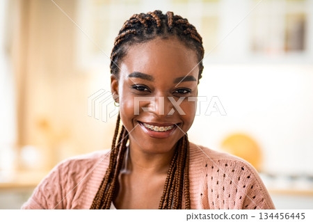 Closeup Portrait Of Smiling African American Female With Braided Hair And Dental Braces Posing At Home Interior, Happy Millennial Black Lady Sitting At Table In Kitchen And Looking At Camera Closeup Portrait Of Smiling African American Female With Braided Hair And Dental Braces Posing At Home Interior, Happy Millennial Black Lady Sitting At Table In Kitchen And Looking At Camera 134456445